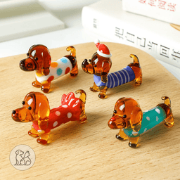 A collection of various mini hand-blown glass dachshund dog figurines, featuring colorful patterns with polka dots, stripes, and festive holiday hats, shown with size measurements on a desk setup.