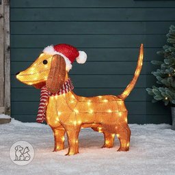 Pre-lit 2.3ft Christmas lighted dachshund wearing a red Santa hat and striped scarf standing on snow in an outdoor yard with warm glowing LED lights.