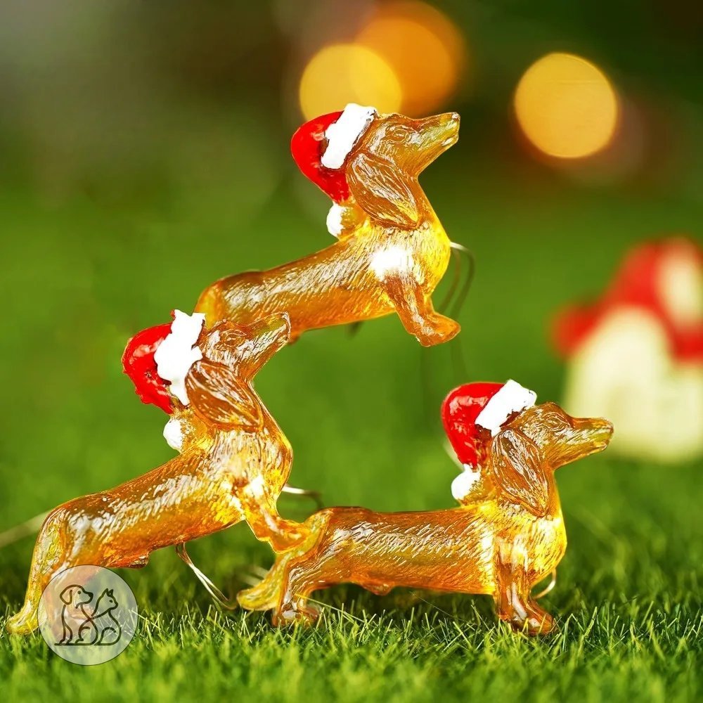 Warm glowing Christmas string lights shaped like miniature golden dachshund dogs wearing red Santa hats lit up on green grass.
