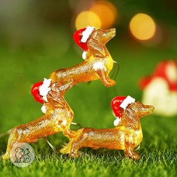 Warm glowing Christmas string lights shaped like miniature golden dachshund dogs wearing red Santa hats lit up on green grass.