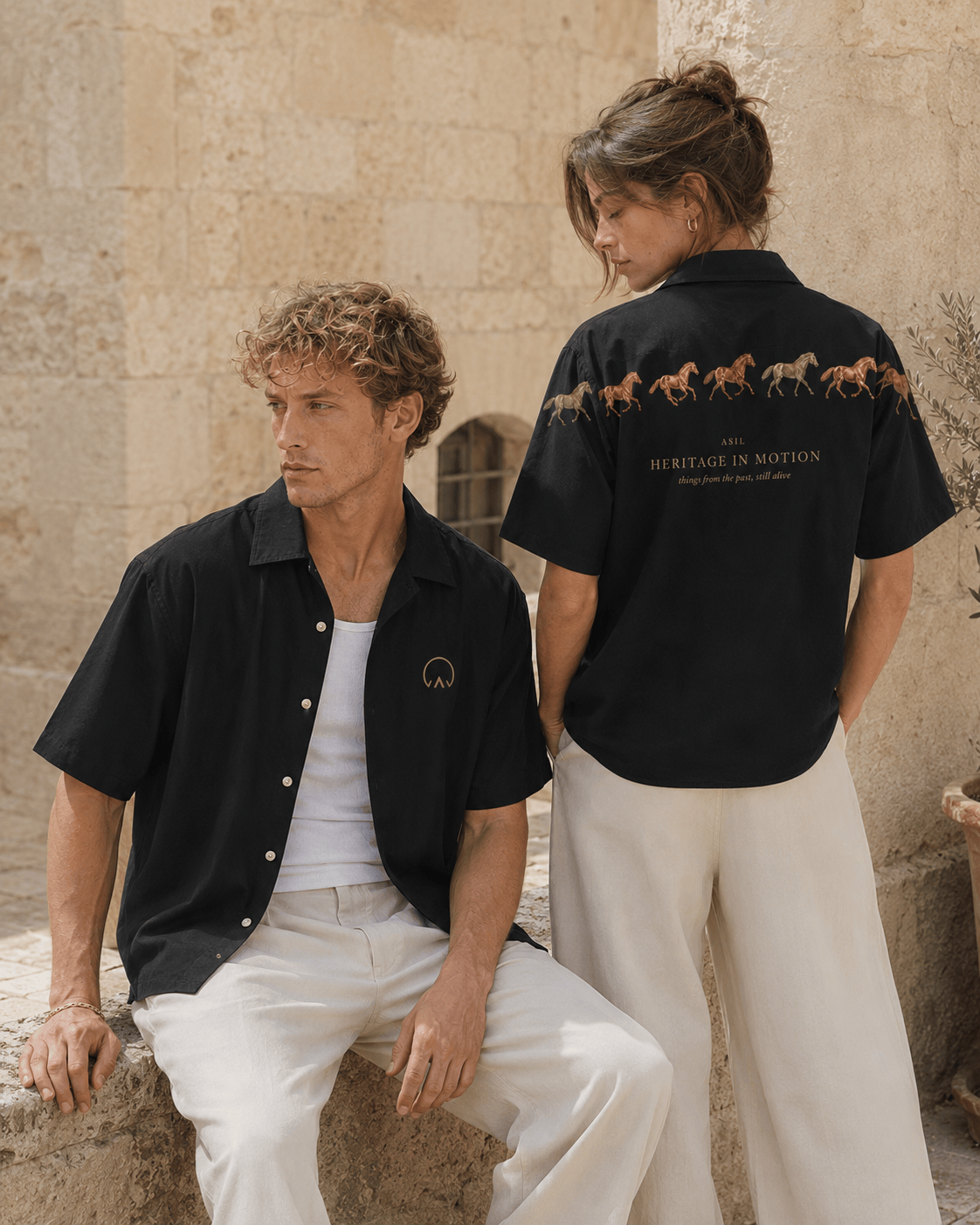 Male and female models wearing the black Asil short-sleeve shirts, with the front chest emblem and Heritage in Motion horse artwork showcased in a warm heritage stone courtyard.