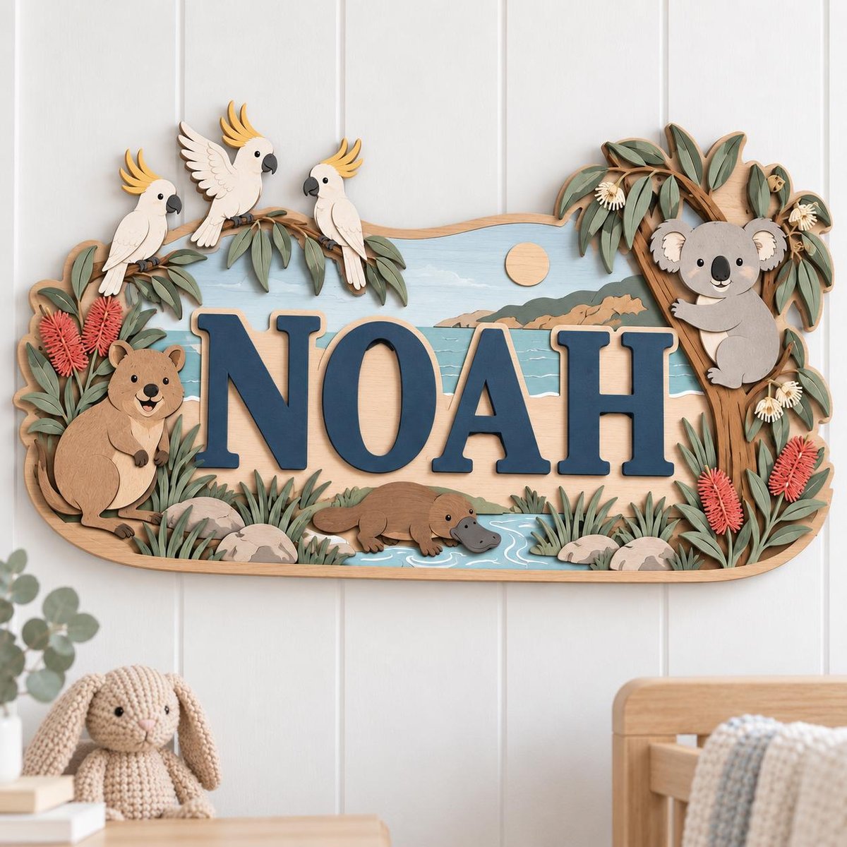 May include: A layered wooden wall sign featuring the name NOAH in large dark blue letters. The scene depicts Australian wildlife including three white cockatoos, a koala in a tree, a quokka, and a platypus, surrounded by eucalyptus leaves and red flowers.
