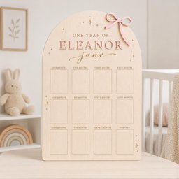 May include: An arched wooden milestone board featuring the text ONE YEAR OF ELEANOR JANE, a pink bow accent, and twelve rectangular photo frames labeled from one month to one year, decorated with small engraved star symbols on the light wood surface.