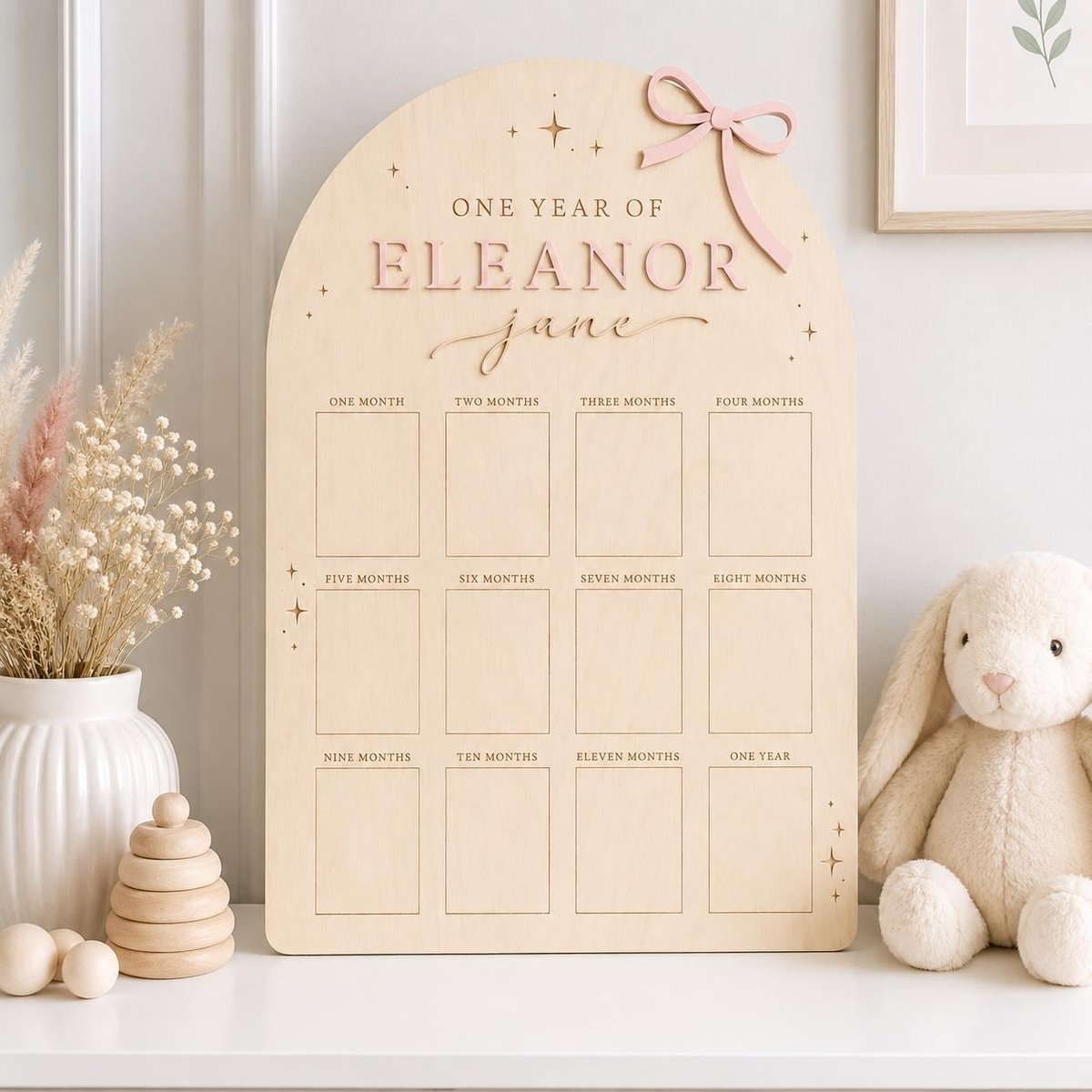 May include: An arched wooden milestone board featuring the text ONE YEAR OF ELEANOR JANE, a pink bow accent, and twelve rectangular photo frames labeled from one month to one year, decorated with small star icons engraved on the light wood surface.