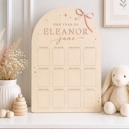 May include: An arched wooden milestone board featuring the text ONE YEAR OF ELEANOR JANE, a pink bow accent, and twelve rectangular photo frames labeled from one month to one year, decorated with small star icons engraved on the light wood surface.
