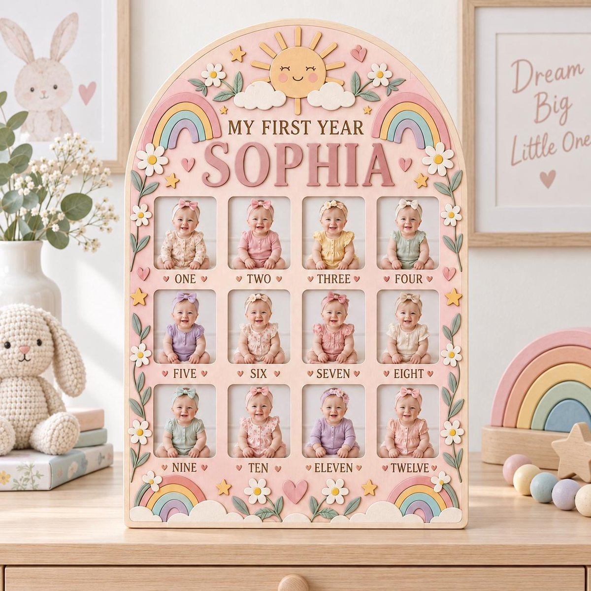 May include: A light pink arched wooden photo frame titled MY FIRST YEAR SOPHIA, featuring twelve rectangular slots for monthly baby photos. The frame is decorated with pastel rainbows, a smiling sun, white daisies, and small hearts and stars.