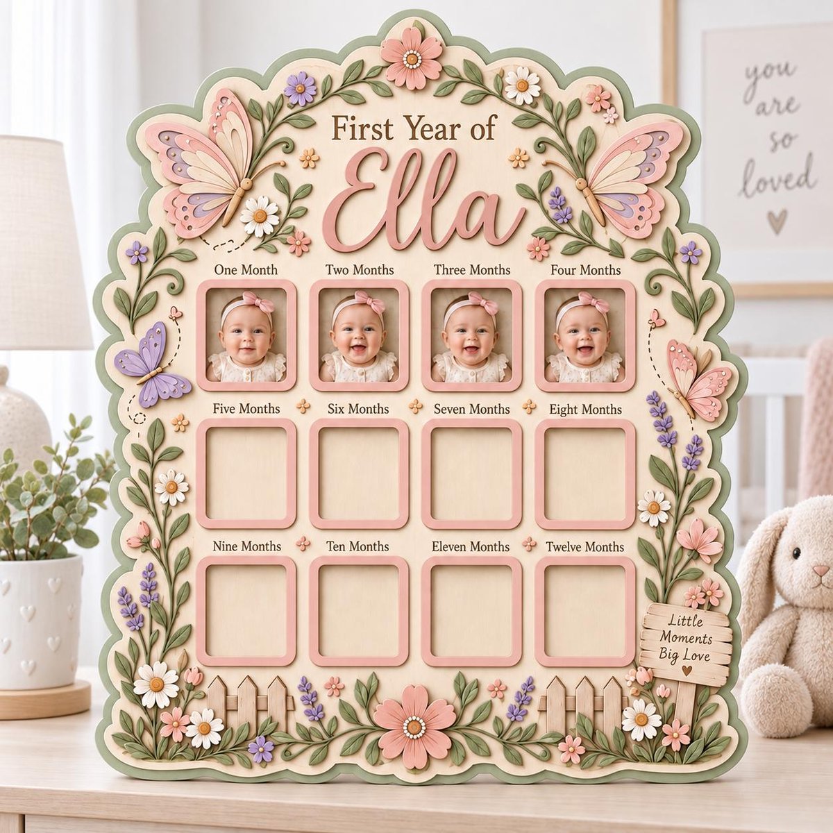 May include: A wooden baby milestone photo board titled First Year of Ella with twelve square frames. The board features pastel-colored butterflies, flowers, and vines. It includes text labels for each month and a small sign reading Little Moments Big Love.