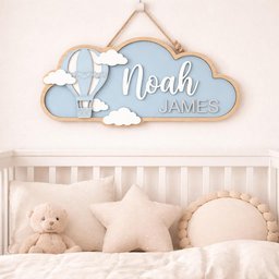 May include: A wooden cloud-shaped sign with the name "Noah JAMES" in white lettering. The sign features a light blue background, a hot air balloon, and white clouds. It hangs above a crib with pillows and a teddy bear.