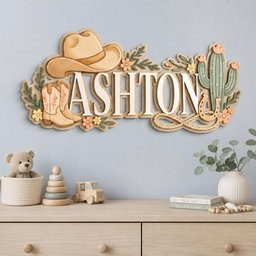 May include: A decorative wooden wall sign featuring the name ASHTON in large white letters. The design includes a tan cowboy hat, a pair of tan cowboy boots, a green cactus, a rope lasso, and small orange and yellow flowers with green leaves.
