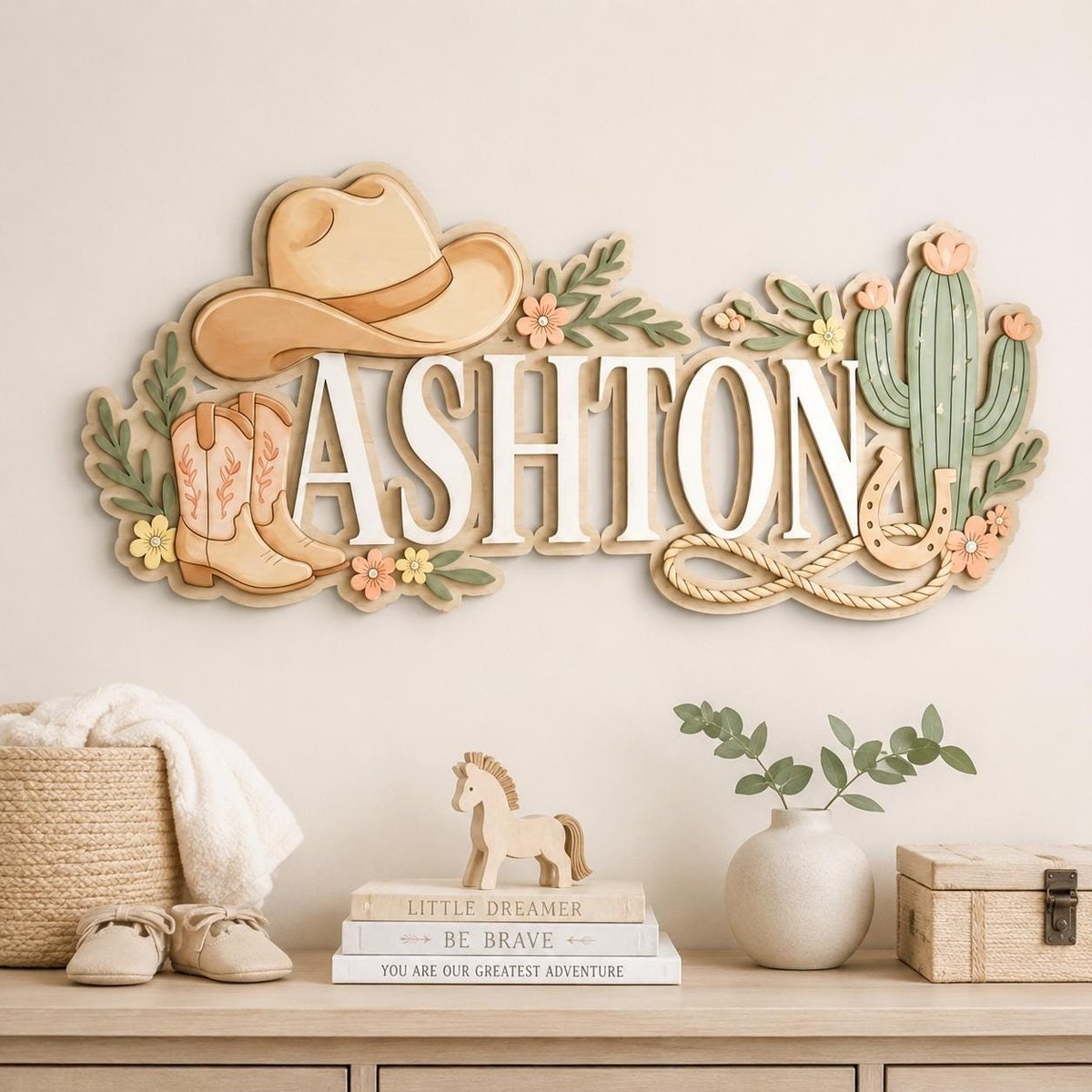 May include: A layered wooden wall sign featuring the name ASHTON in white capital letters. The design includes a tan cowboy hat, a pair of tan cowboy boots, a green cactus, a horseshoe, and small floral accents in shades of pink and yellow.