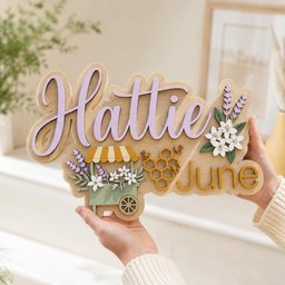 May include: A layered wooden sign featuring the name Hattie in large purple script and the word June in gold block letters. The design includes a green cart with white flowers and lavender, a gold honeycomb pattern with bees, and floral accents.
