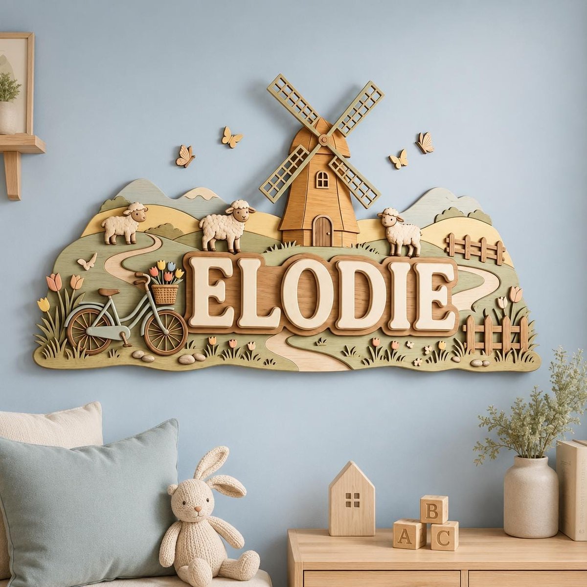 May include: A layered wooden wall sign featuring the name ELODIE in large capital letters. The design includes a central windmill, three sheep, a bicycle with a flower basket, butterflies, rolling hills, and a fence in shades of green and tan.