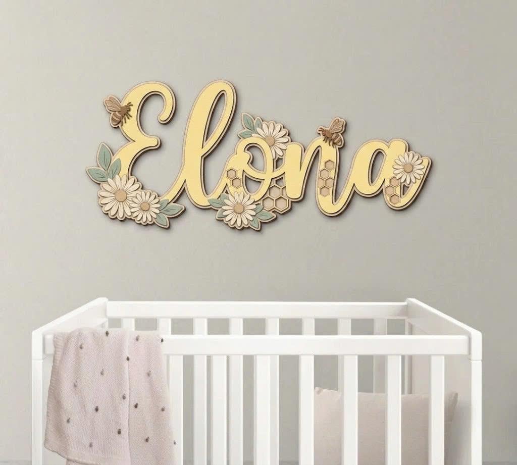 May include: A wooden wall sign featuring the name Elona in cursive script, decorated with layered daisies, green leaves, honeycombs, and small bees. The sign is rendered in shades of yellow, cream, and sage green, mounted above a white crib.