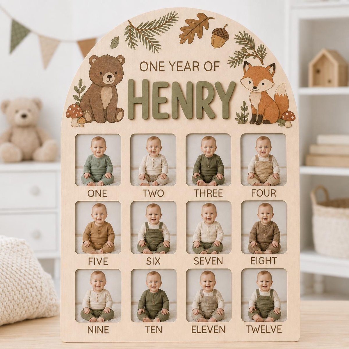 May include: A light-colored wooden arch-shaped photo board featuring the text ONE YEAR OF HENRY, a bear, a fox, and forest elements. It includes twelve square cutouts for photos labeled ONE through TWELVE, displaying a baby in each frame.